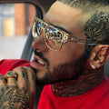 Man with tattoos wearing gold sunglasses and a red shirt, sitting inside a vehicle.