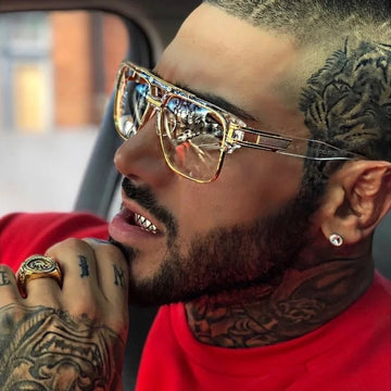 Man with tattoos wearing gold sunglasses and a red shirt, sitting inside a vehicle.