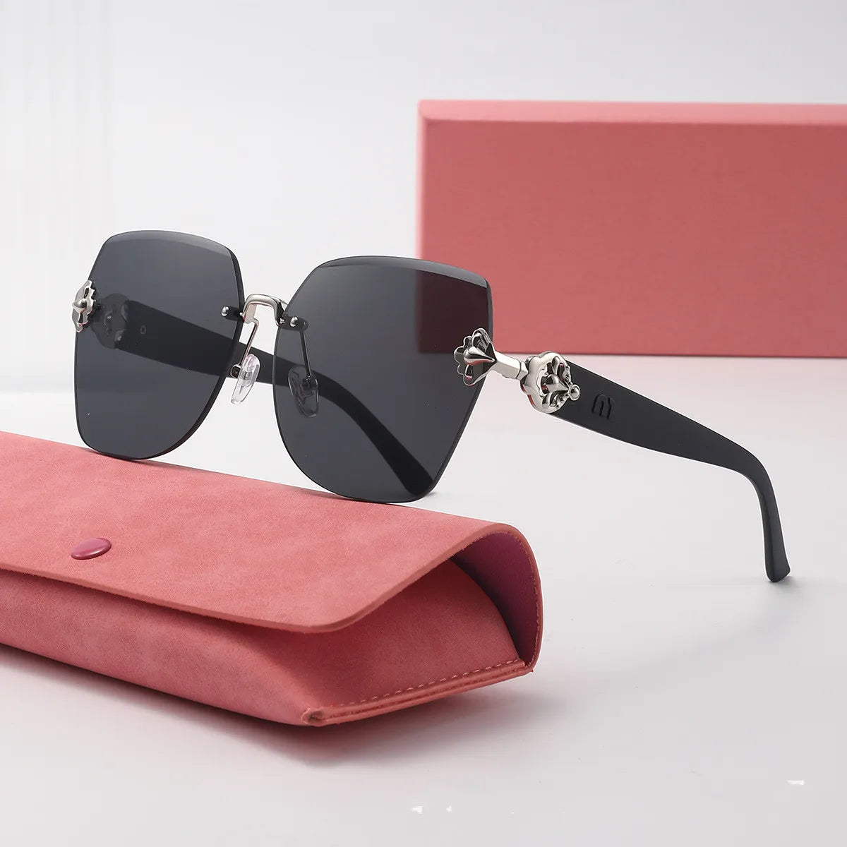 Black sunglasses with decorative arms on a pink case and box against a light gray background